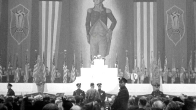 A photograph of the 1939 nazi "German American Bund" rally at Madison Square Garden. A crowd of nazis stands in front of a stage. Across the stage is a row of U.S. flags and centered is a towering portrait George Washington, flanked by U.S. and nazi flags.