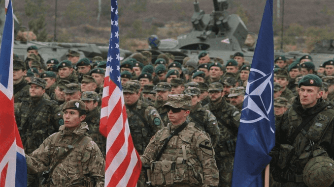 Photo of deployed soldiers standing at attention with US, NATO, and UK flags.