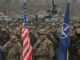 Photo of deployed soldiers standing at attention with US, NATO, and UK flags.