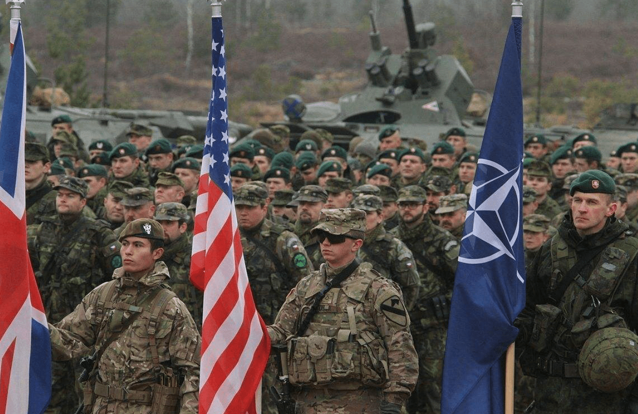 Photo of deployed soldiers standing at attention with US, NATO, and UK flags.