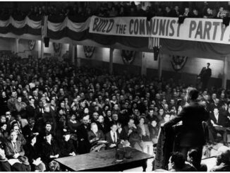 Black and white photograph of a full auditorium from the rostrum; a sign reads BUILD THE COMMUNIST PARTY!