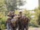 Photograph of men in fatigues and combat helmets being held hostage from behind by men in armored vests and sunglasses pointing pistols at the napes of their necks.