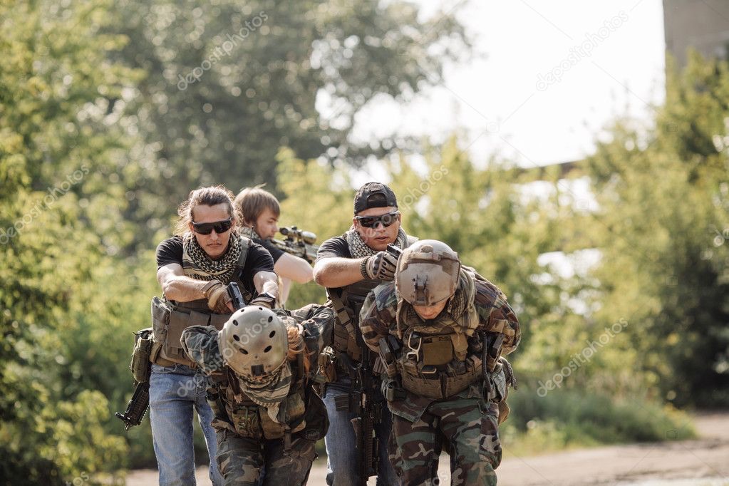 Photograph of men in fatigues and combat helmets being held hostage from behind by men in armored vests and sunglasses pointing pistols at the napes of their necks.