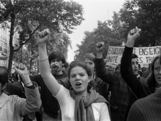 Black and white photograph featuring a woman in a white sweater raising a power fist while marching in the May '68 protests on a tree-lined street