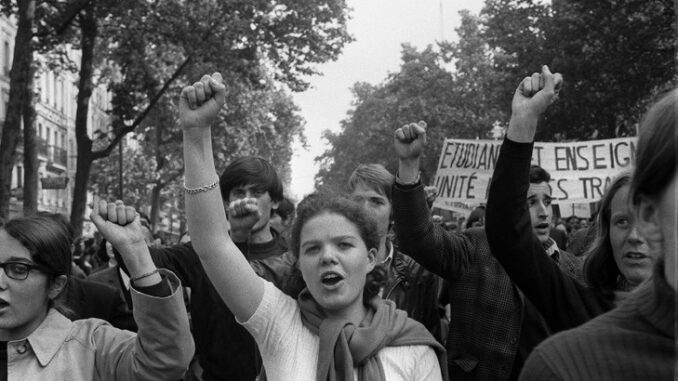 Black and white photograph featuring a woman in a white sweater raising a power fist while marching in the May '68 protests on a tree-lined street